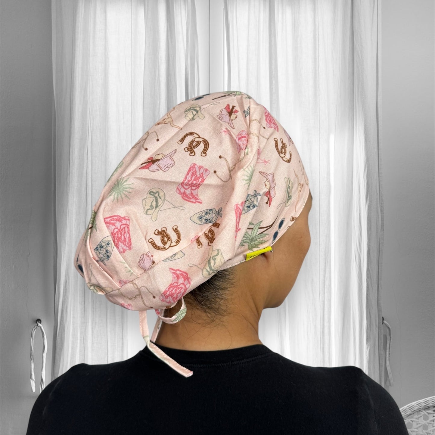 A person wearing a light-colored scrub cap with a playful pattern. The cap features various small illustrations, including cowboy hats, boots, horseshoes, and other western-themed designs.