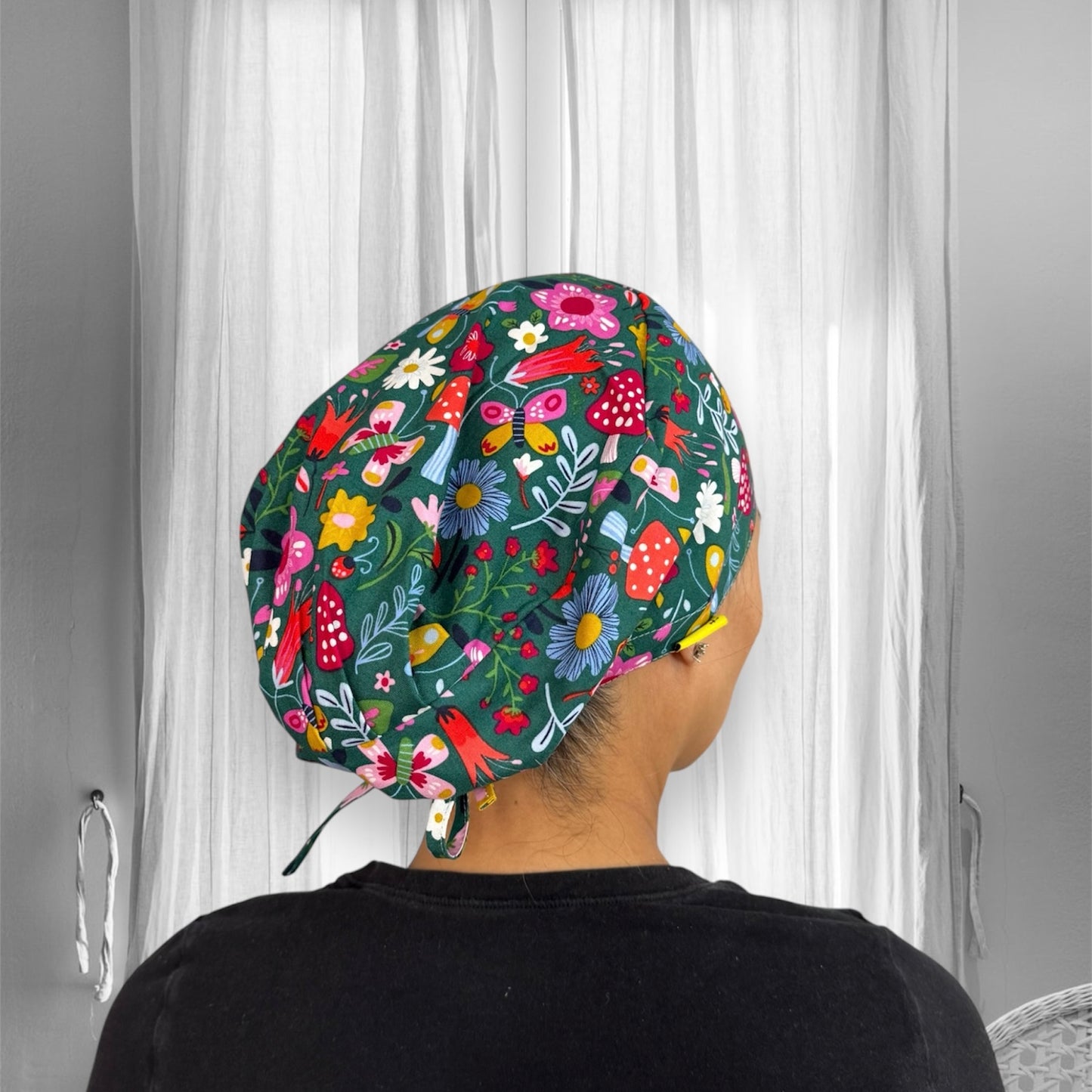 A person is wearing a scrub cap featuring a vibrant floral pattern with various colorful flowers, mushrooms, and leaves on a dark green background.