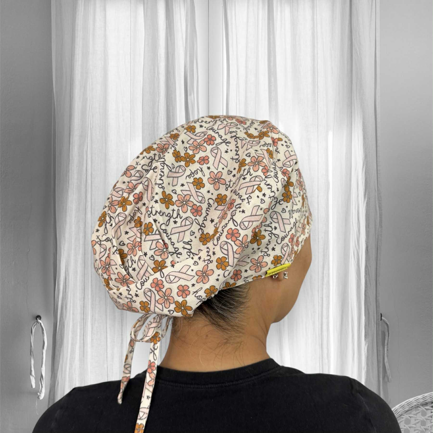 A person wearing a scrub cap. The cap features a pattern of pink awareness ribbons, flowers, and words like "strength," "courage," and "hope" in a handwritten style.