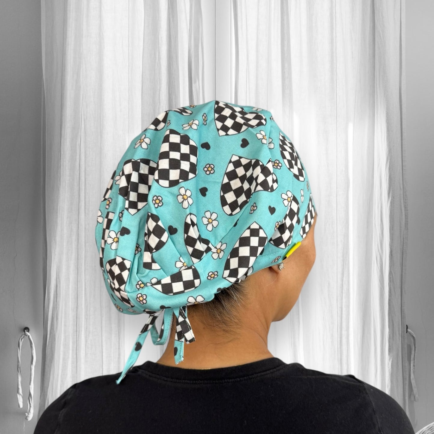 A uniquely patterned scrub cap featuring a light blue base with black and white checkered hearts, small white daisies, and tiny black hearts.