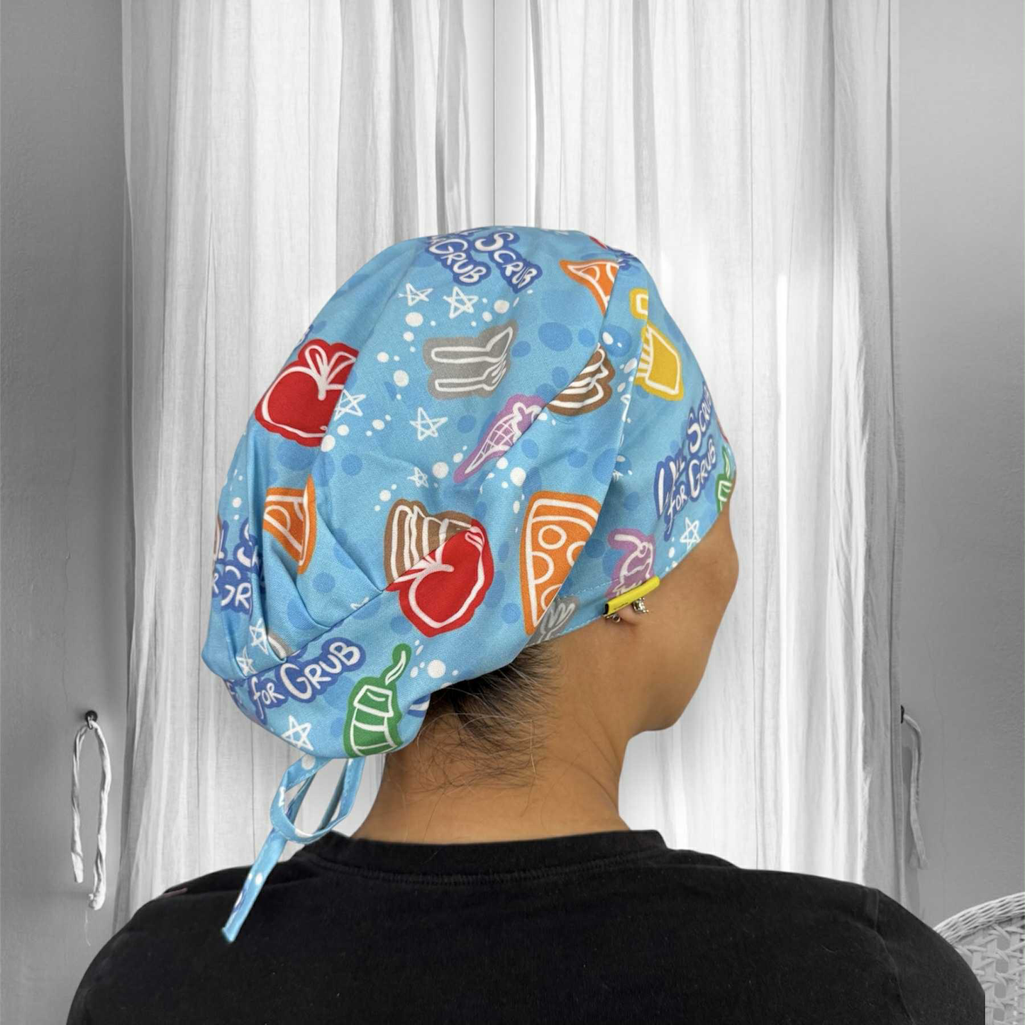 A person wearing a brightly colored scrub cap. The cap is blue with playful, vibrant illustrations featuring various food items like pizza slices, ice cream cones, cupcakes, apples, and drinks. The words "Scrub for Grub" are visible as part of the design.