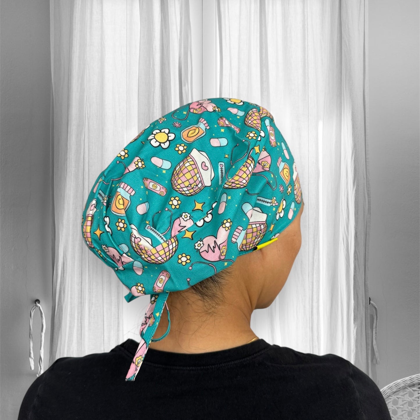 A person wearing a colorful scrub cap with a playful design. The cap features illustrations of various beauty and medical-themed items on a teal background.