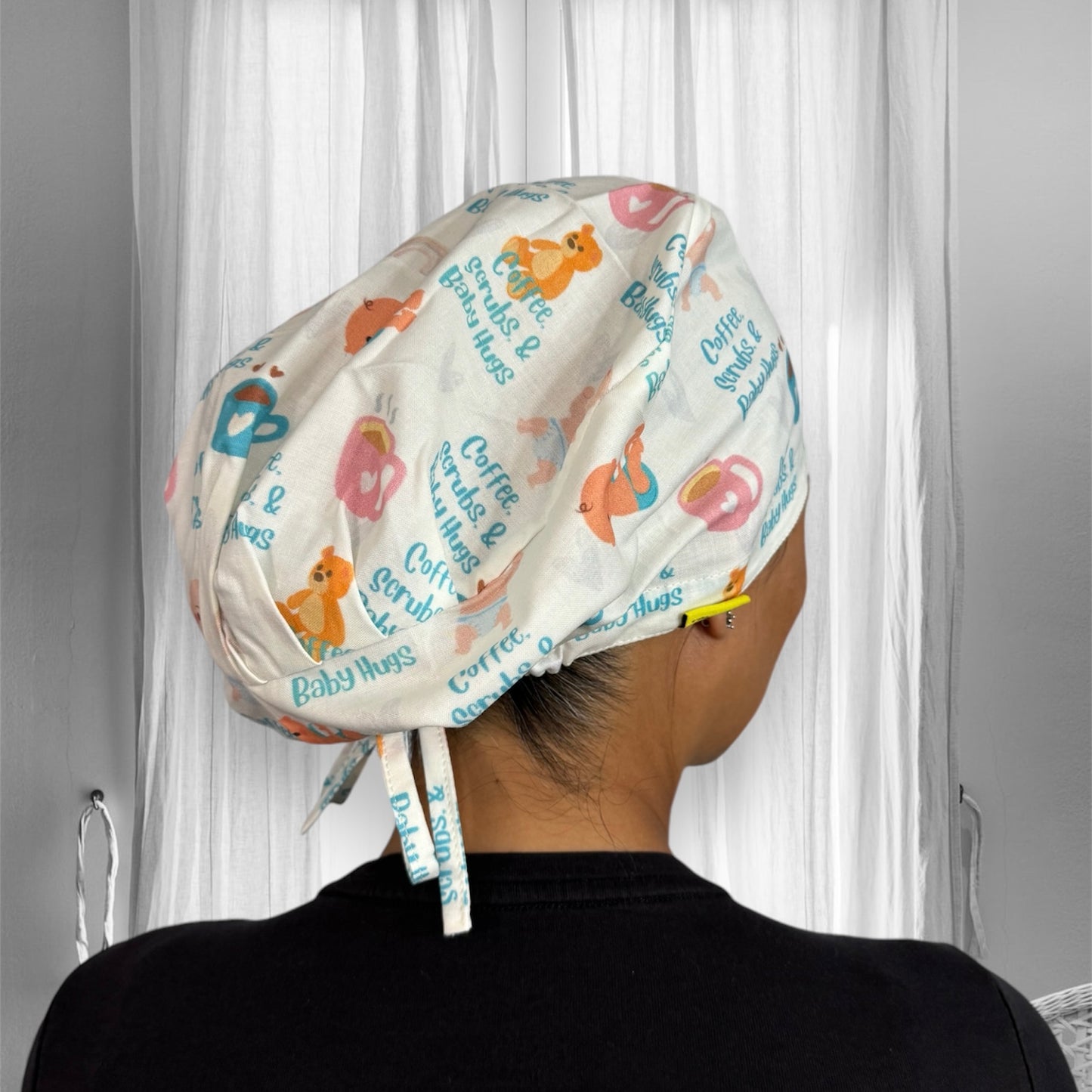 A person wearing a scrub cap with a playful print featuring coffee cups, teddy bears, and text that reads "Coffee, Scrubs & Baby Hugs." The cap's background is white, and the illustrations are in pastel colors like pink, blue, and orange.
