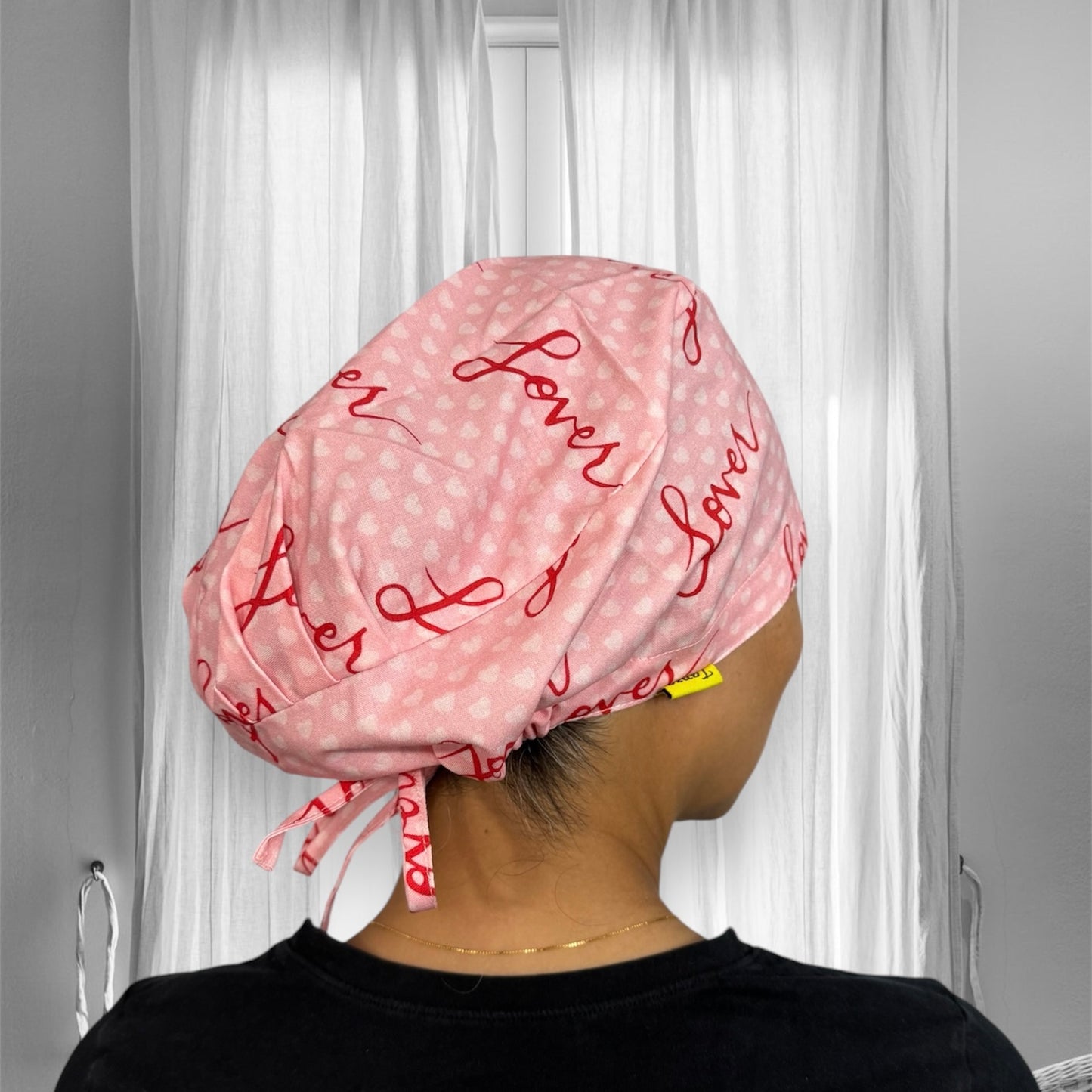 A person is wearing a pink scrub cap with red script writing that reads "lover." The cap features a subtle pattern of small hearts and has ties at the back.