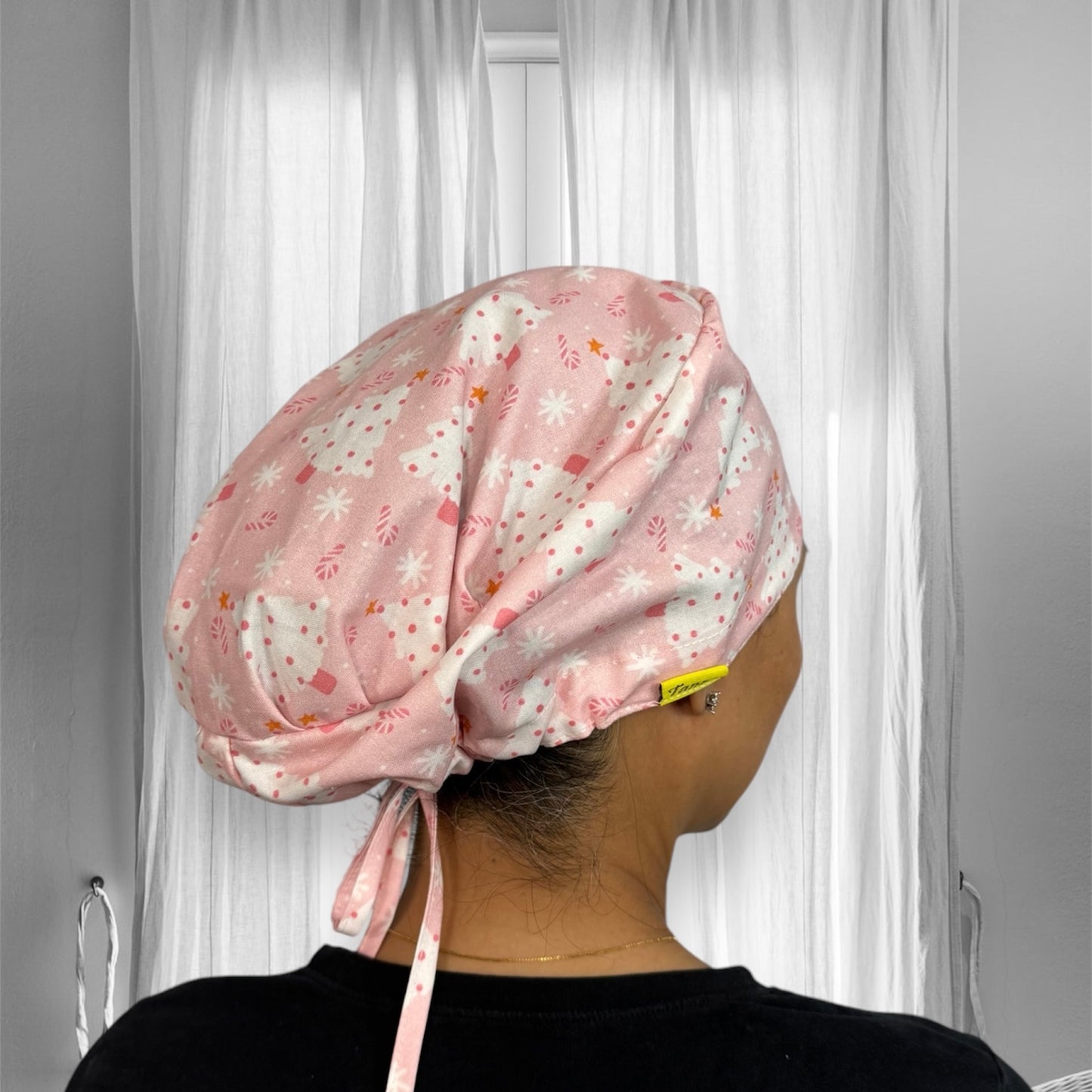 A person wearing a pink scrub cap with a festive pattern featuring white Christmas trees and pink stars. The cap is tied securely at the back of the head, and the material appears soft and comfortable.