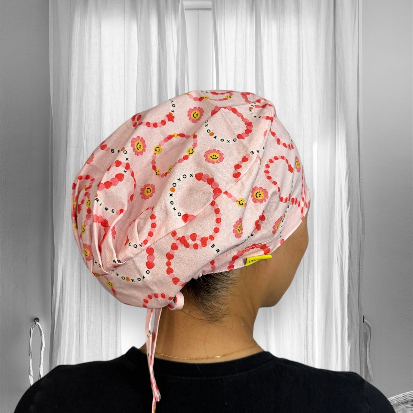 A person wearing a scrub cap with a pink background adorned with patterns of red dots forming circles and yellow floral designs. The cap also has the words "XOXO", "LOVE", and "MINE" printed in various places.