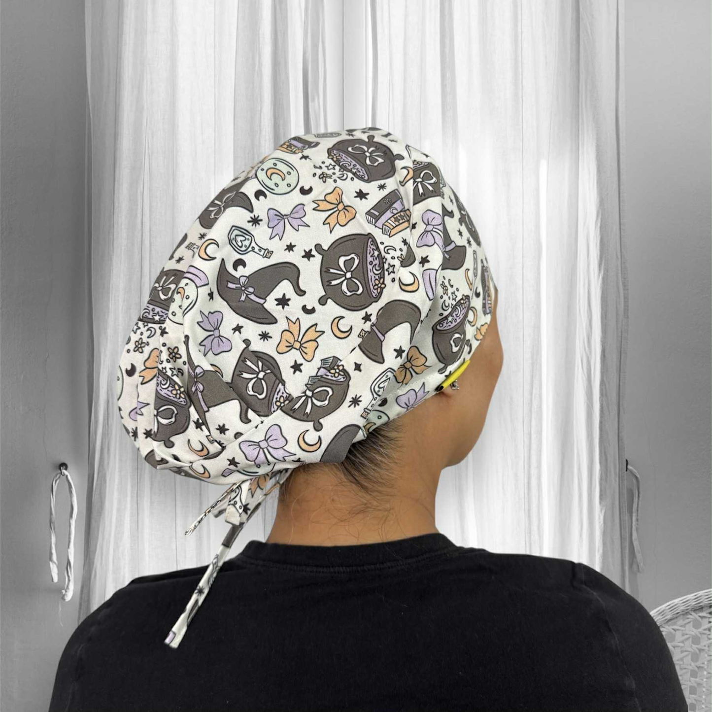 A person wearing a scrub cap with a whimsical pattern. The scrub cap features designs of black cats wearing witch hats, crescent moons, stars, pastel-colored bows, and potion bottles in shades of orange and purple.