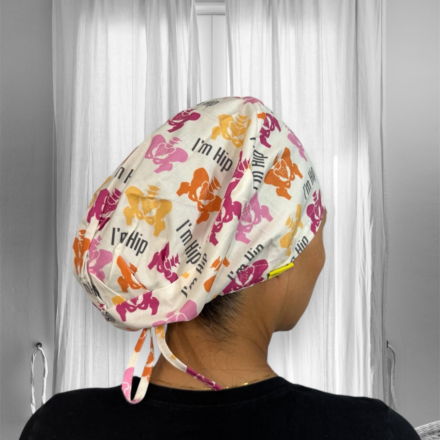 A person wearing a colorful scrub cap with a playful pattern featuring cartoon hippos in pink, orange, and yellow hues. The cap has the phrase "I'm Hip" printed multiple times in a playful font.