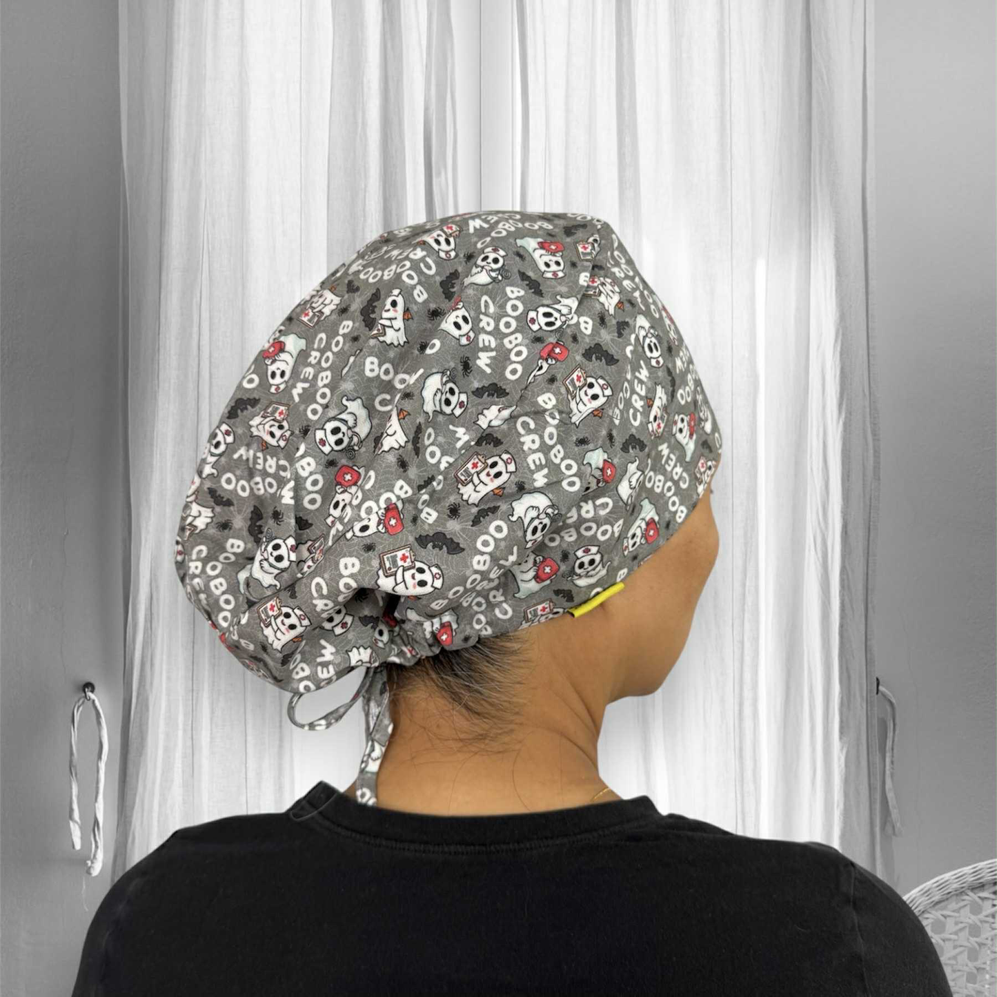 A person wearing a scrub cap with a fun, thematic design. The scrub cap is grey and features a pattern of small white cartoon ghosts and the words "BOO BOO CREW" repeated across it.