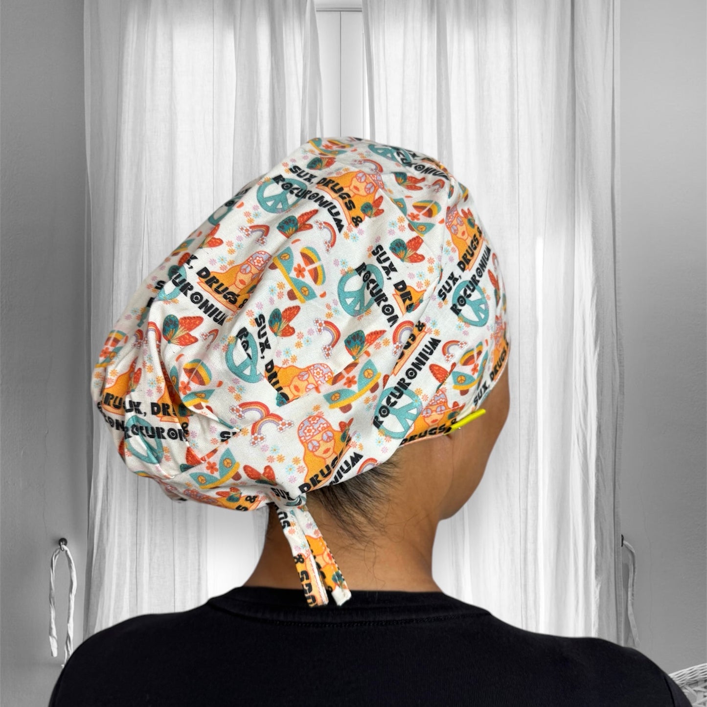 a person wearing a colourful scrub cap with various patterns. The cap features peace symbols, floral designs, and the words "Sux Drugs & Rocuronium" arranged in a whimsical, vibrant style.