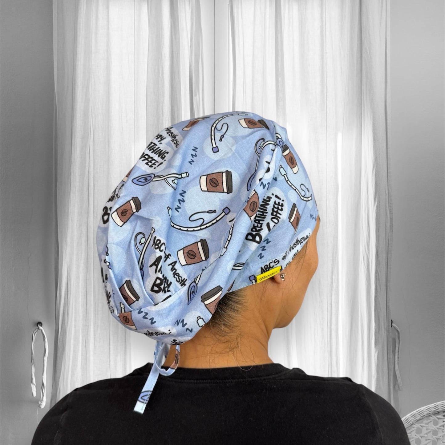 A person wearing a light blue scrub cap with a playful design. The cap features illustrations of coffee cups, stethoscopes, and text such as "Breathing, Coffee," "ABCs of Anesthesia".