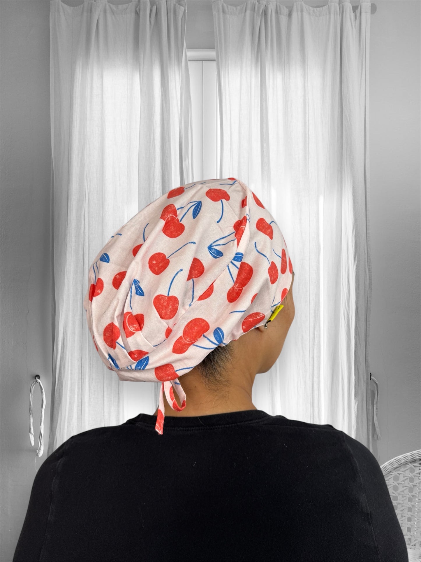 A person wearing a scrub cap with a playful cherry pattern. The scarf is adorned with red cherries and blue stems, tied securely at the back.
