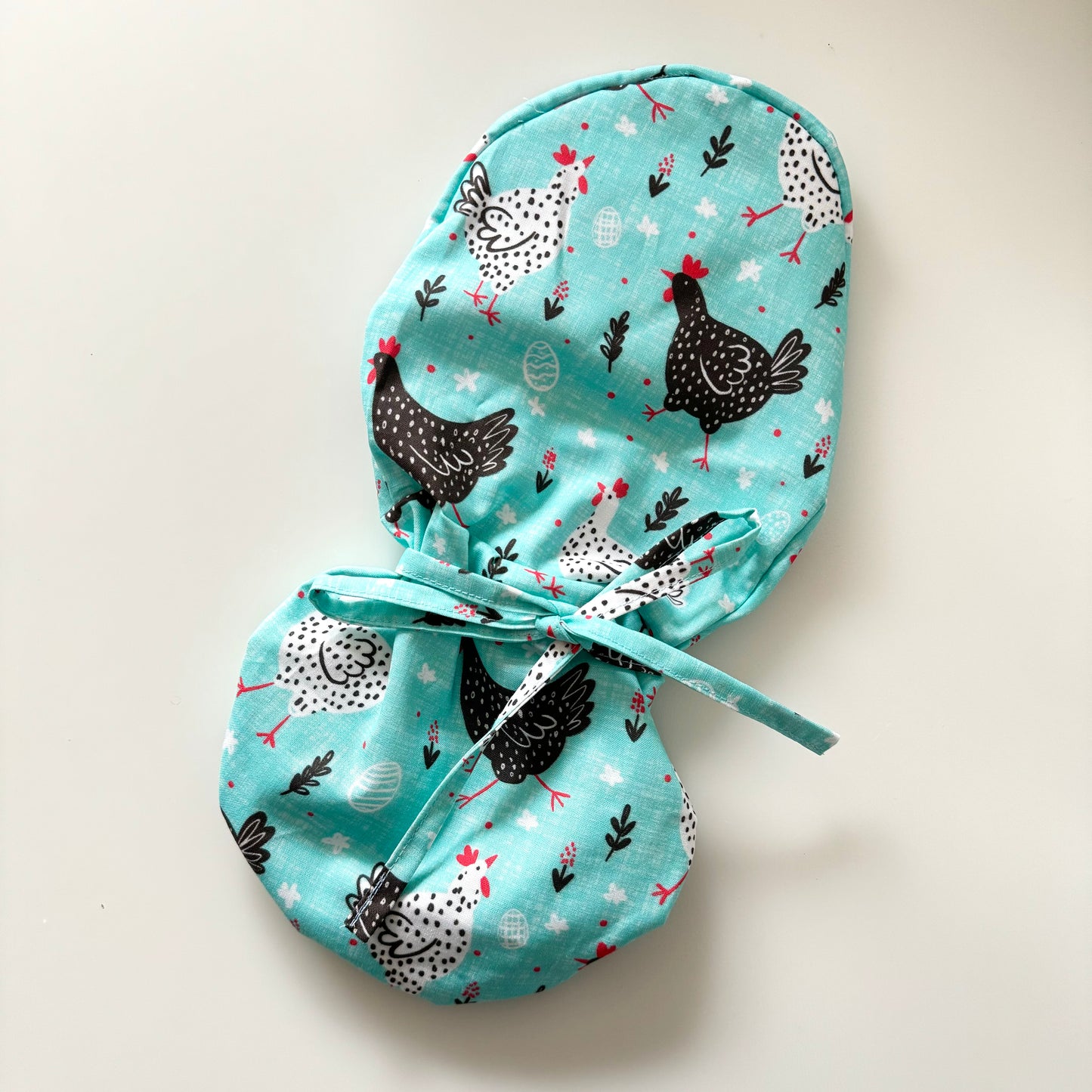 A scrub cap with a playful pattern featuring black and white chickens on a light blue background. The cap has a tie closure and is adorned with small decorative elements like leaves and eggs.
