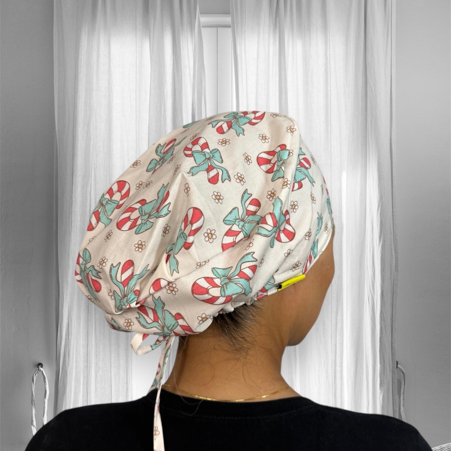 A scrub cap with a festive design featuring candy canes and teal bows. The cap has a soft white background, adorned with small daisy-like flowers scattered throughout.