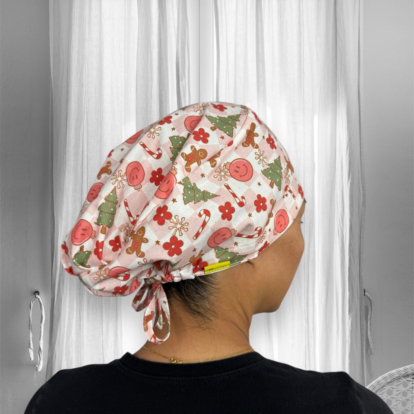 A festive scrub cap featuring a pattern of Christmas-themed designs, including Christmas trees, candy canes, gingerbread men, and red flowers.