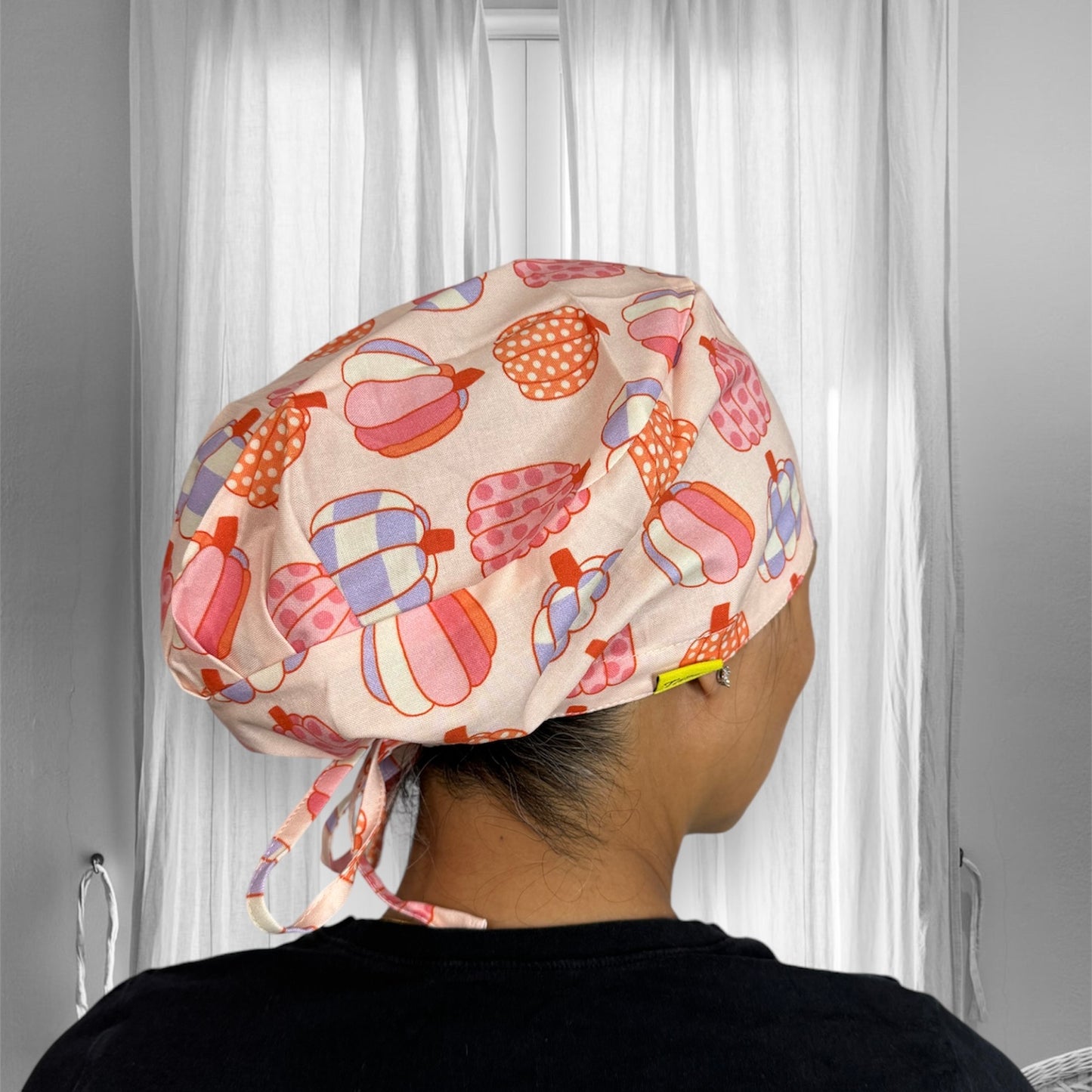 A person is wearing a colorful scrub cap with a tie at the back. The cap features a playful pattern of pastel-colored shapes, including pink, orange, and light blue, against a cream background.