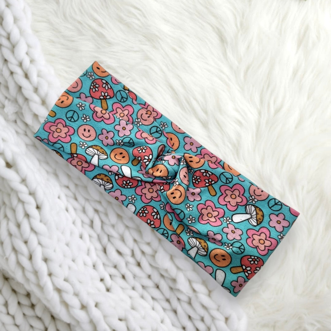A colorful headband featuring a playful design of flowers, smiley faces, peace symbols, and mushrooms on a soft, knitted fabric background.
