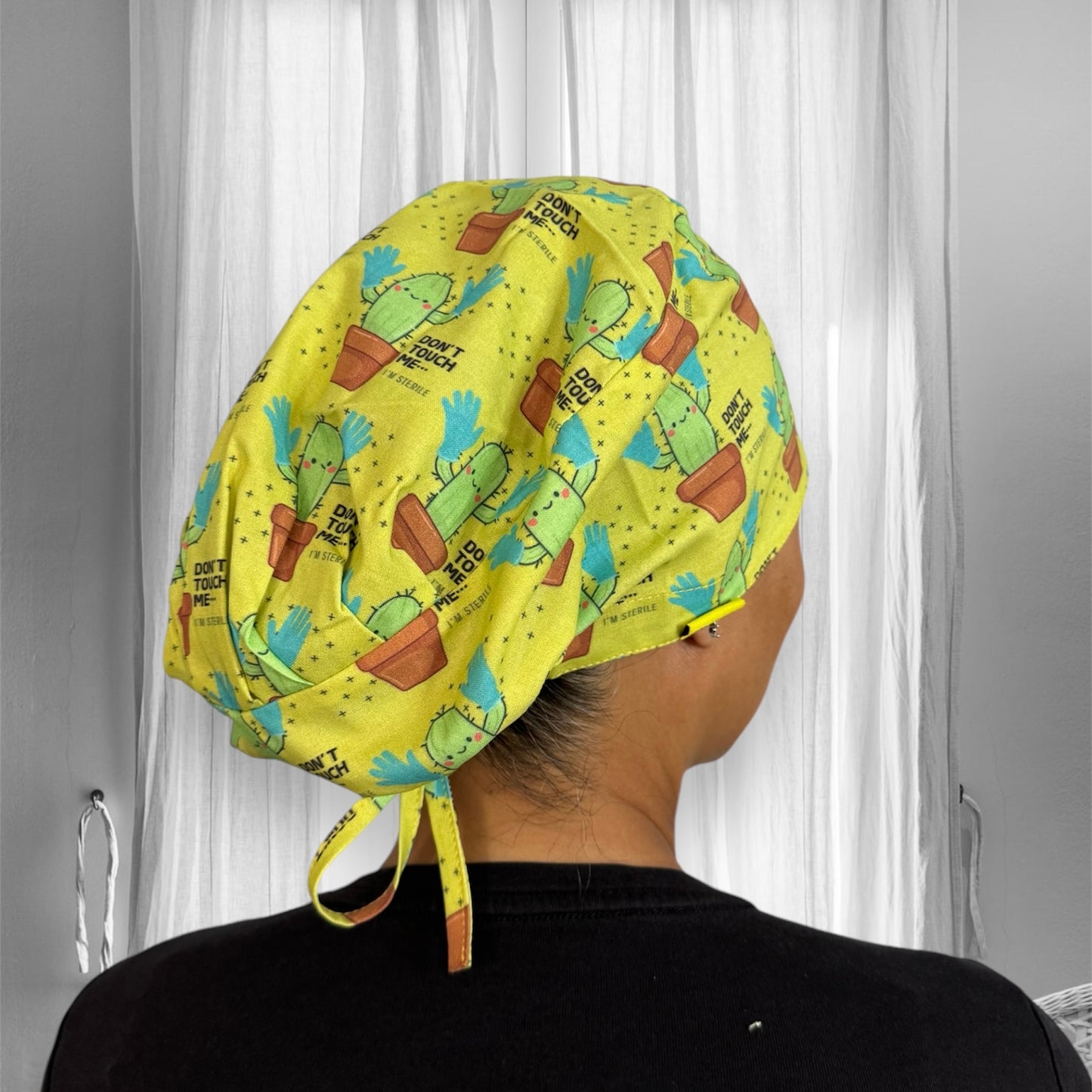 A scrub cap made of yellow fabric adorned with playful cactus illustrations. Each cactus is wearing a pair of blue gloves, and the design includes the phrase "DON'T TOUCH ME!"