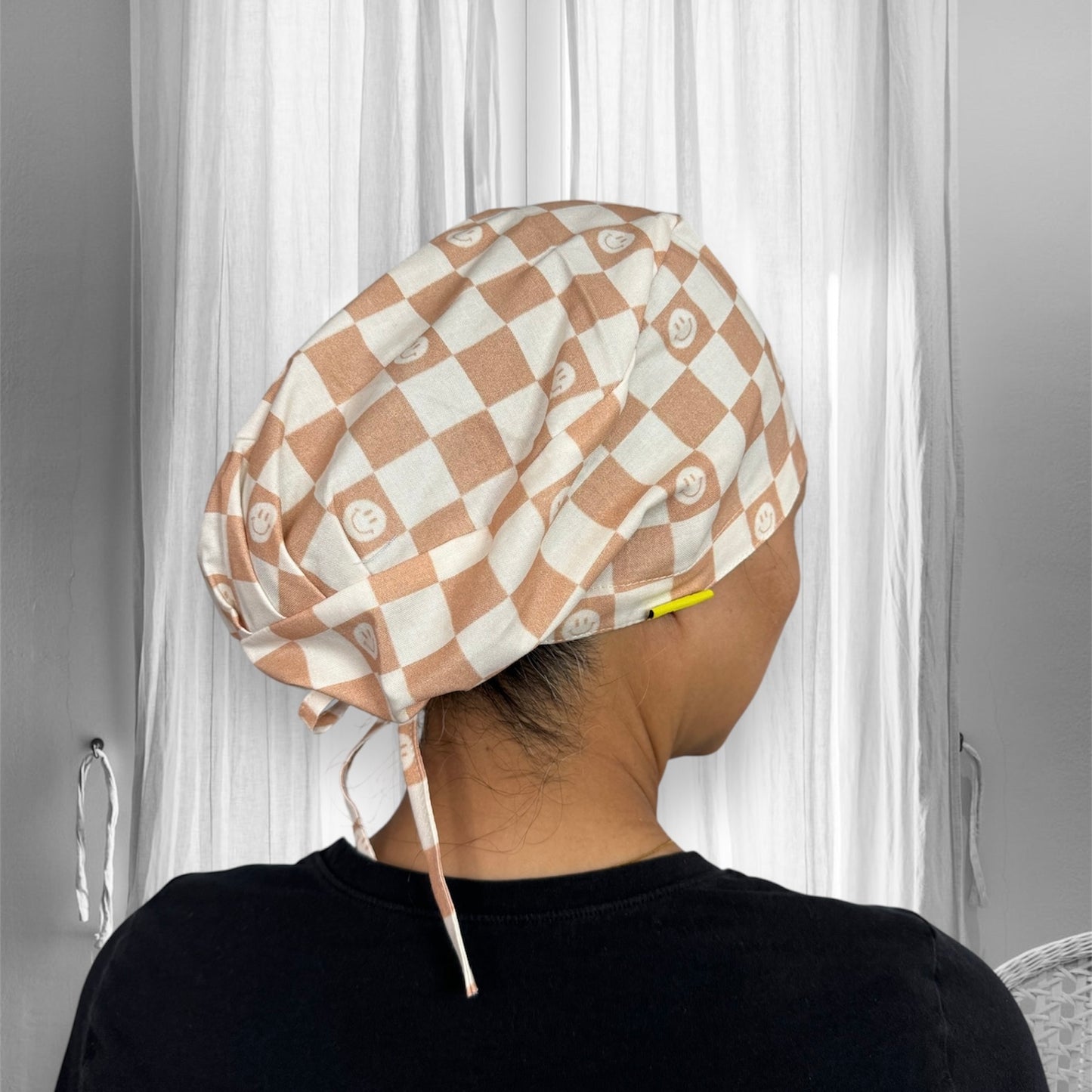 A checkered scrub cap with a beige and white pattern, featuring smiley faces on each square.