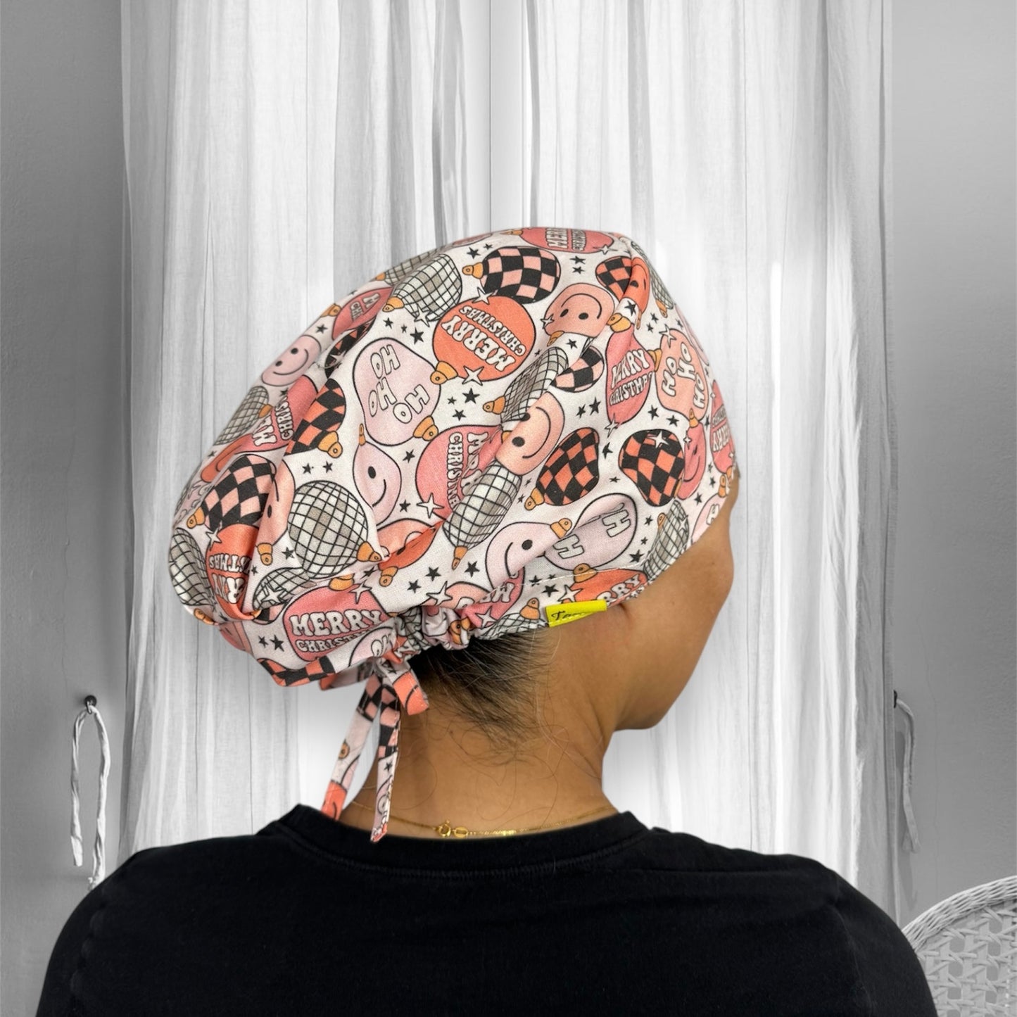 A scrub cap with a festive design. The cap features a variety of Christmas-themed illustrations, including Santa hats, snowflakes, and checkered patterns in shades of pink, red, and black.
