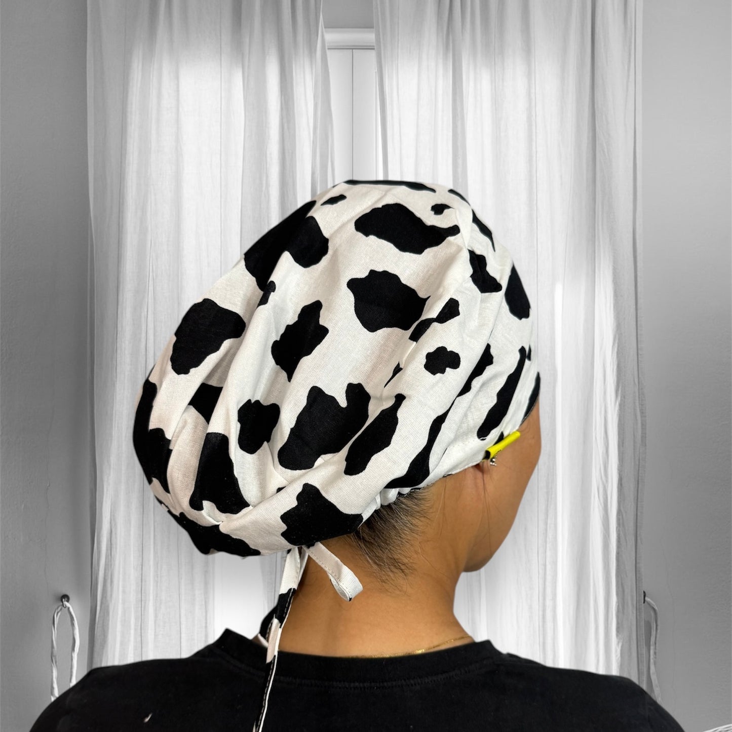 A person wearing a scrub cap with a black and white cow print design. The cap is loose-fitting and tied at the back, with the pattern resembling cowhide patches.