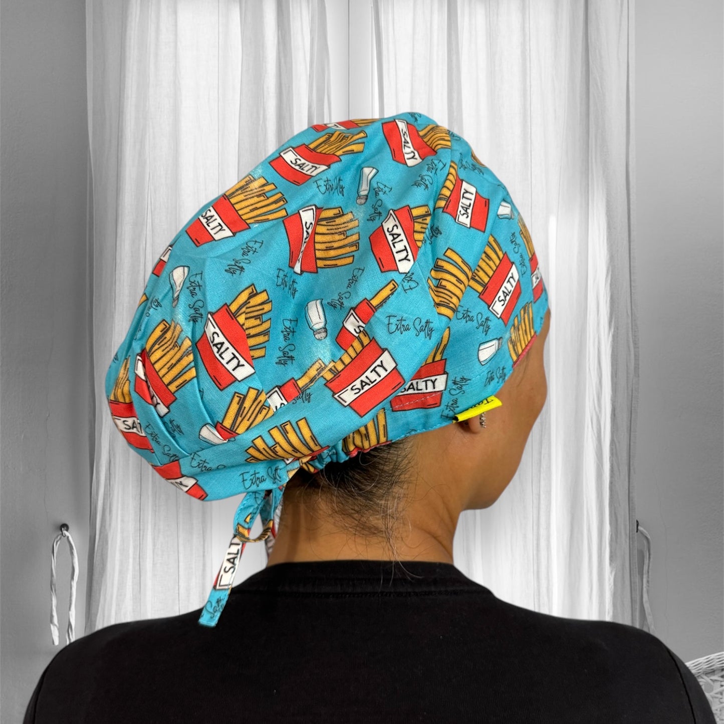 A person wearing a colorful scrub cap that features a playful pattern. The cap is predominantly blue and is adorned with images of French fries and boxes labeled "SALTY."
