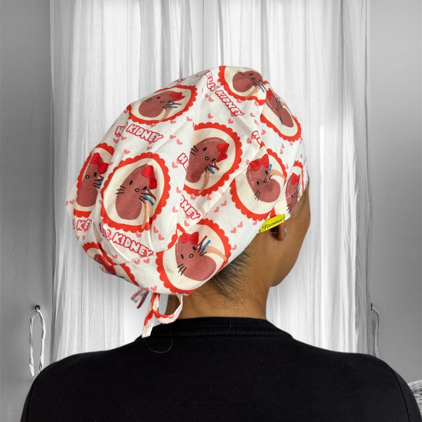 A person wearing a scrub cap with a playful design. The cap features cute illustrations of kidneys with smiling faces, each adorned with a small red bow. The pattern is repeated across the fabric, accompanied by the text "HELLO KIDNEY" surrounded by small red hearts.