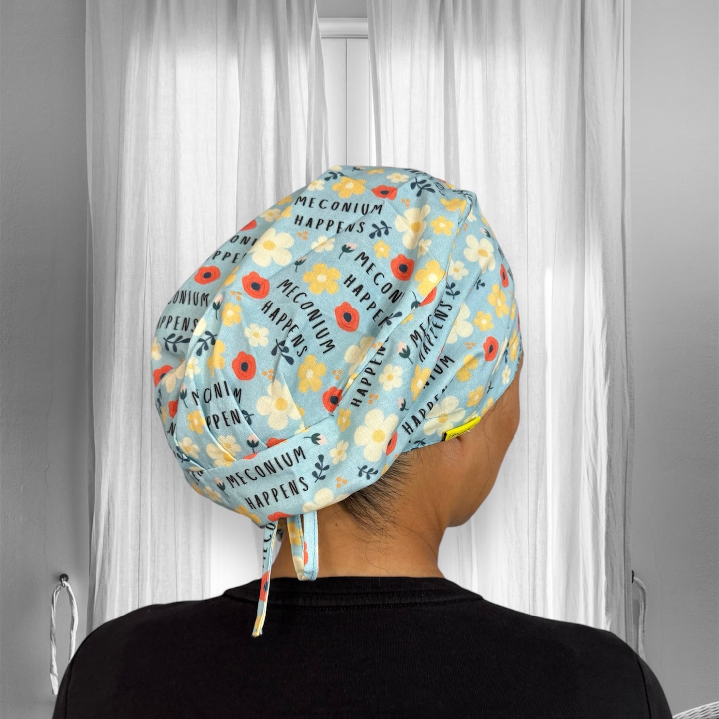 A scrub cap with a light blue background featuring a playful floral pattern, adorned with yellow, white, and orange flowers. The text "Meconium Happens" is repeated across the fabric.