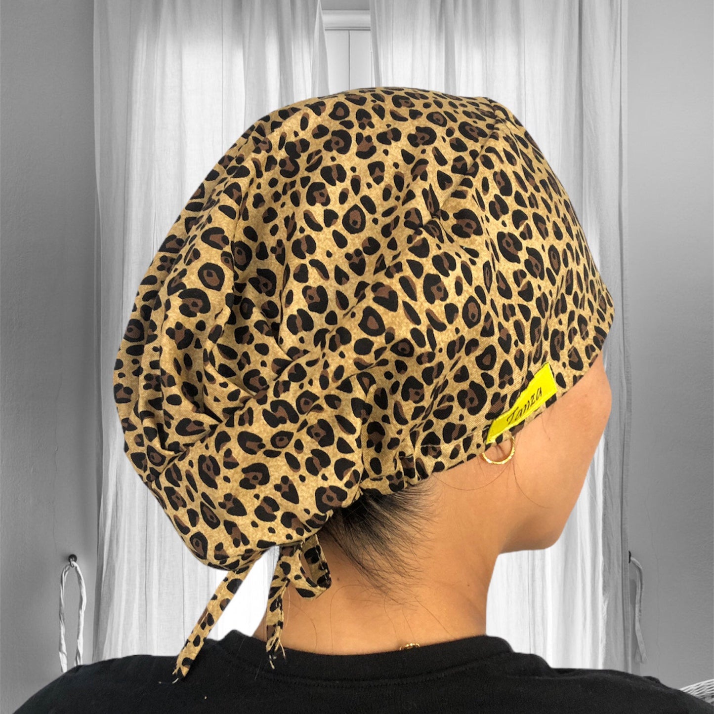 A cheetah print scrub cap with a tie at the back. The cap features a bold, spotted pattern in shades of brown and black.
