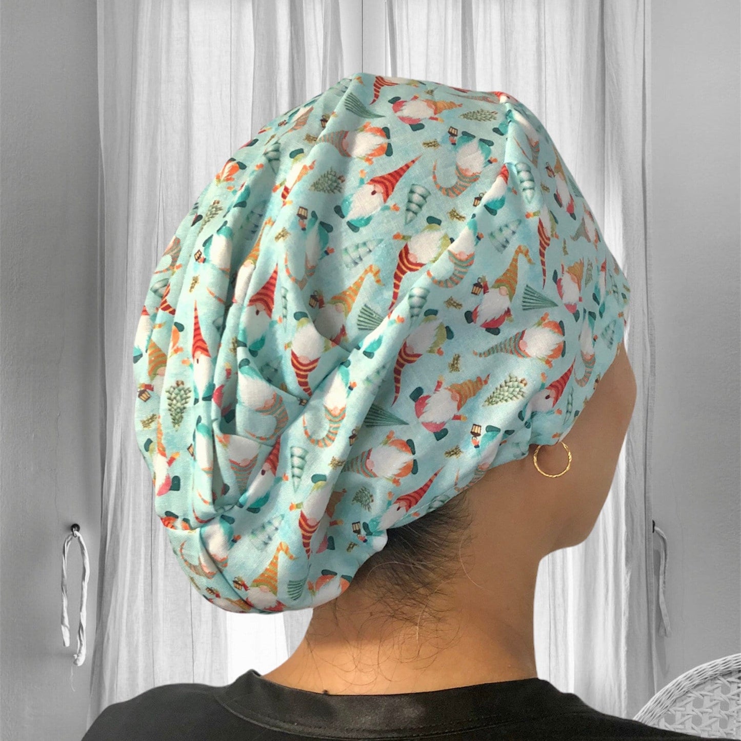 A person wearing a stylish scrub cap adorned with a whimsical pattern of gnomes and festive elements. The cap is in a light blue color, with the gnomes sporting red and white hats, adding a playful and festive touch.