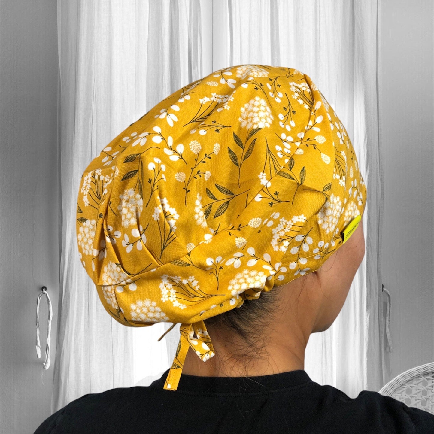 A person wearing a vibrant yellow scrub cap with a floral print design. The cap features a pattern of white flowers and green leaves, and is tied at the back.