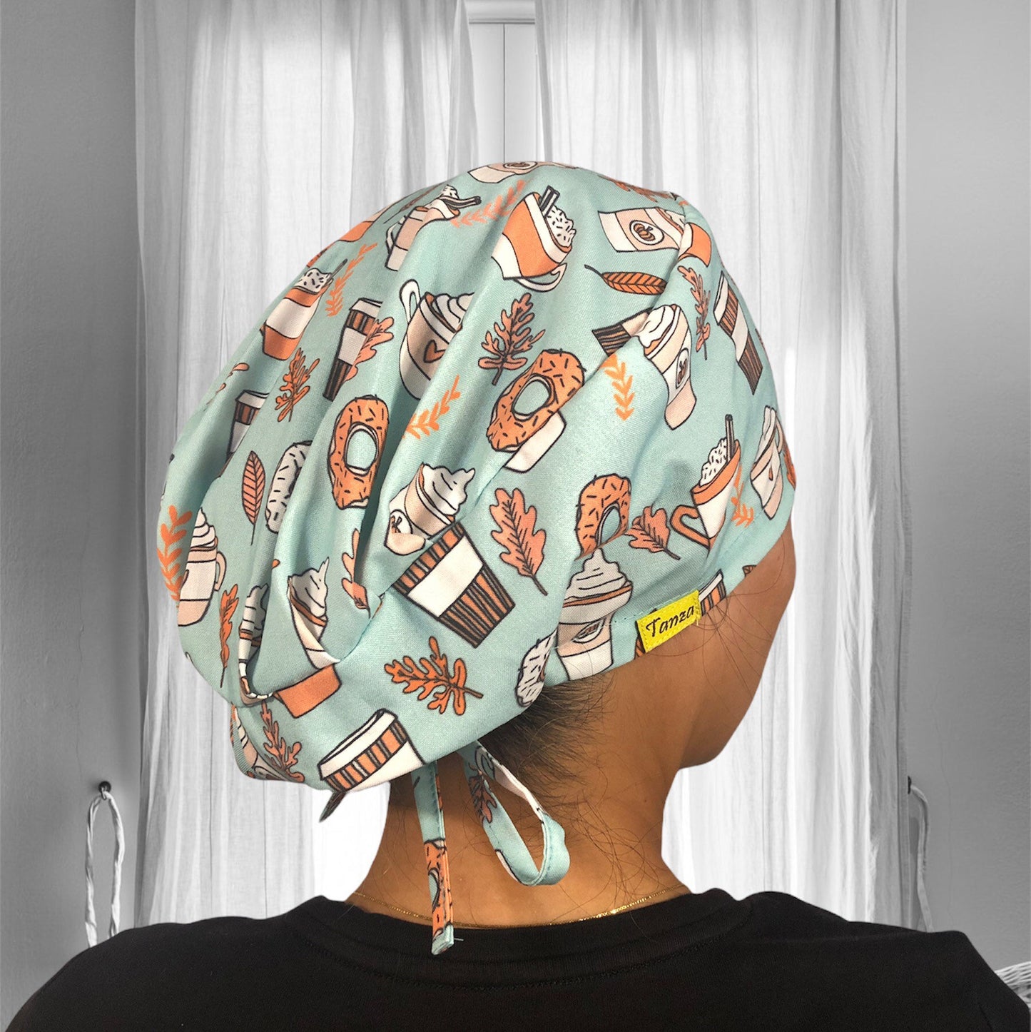A person wearing a scrub cap with a playful pattern. The cap is adorned with illustrations of various coffee and pastry items, like cups of coffee, donuts, and croissants. The fabric's background is a soft blue, and the illustrations are in shades of orange and white.