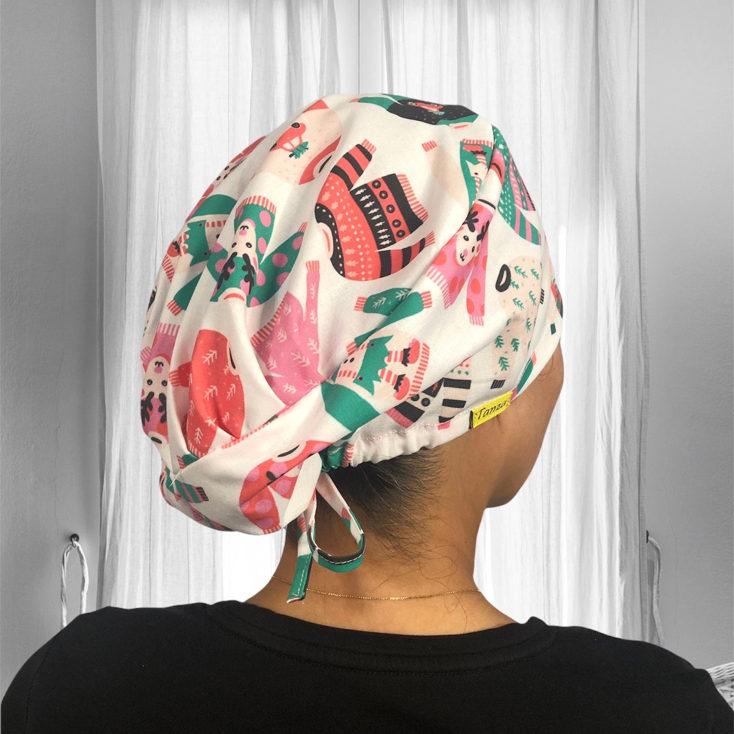A colorful scrub cap featuring a festive design with cartoon characters wearing winter-themed sweaters and hats, set against a white background.