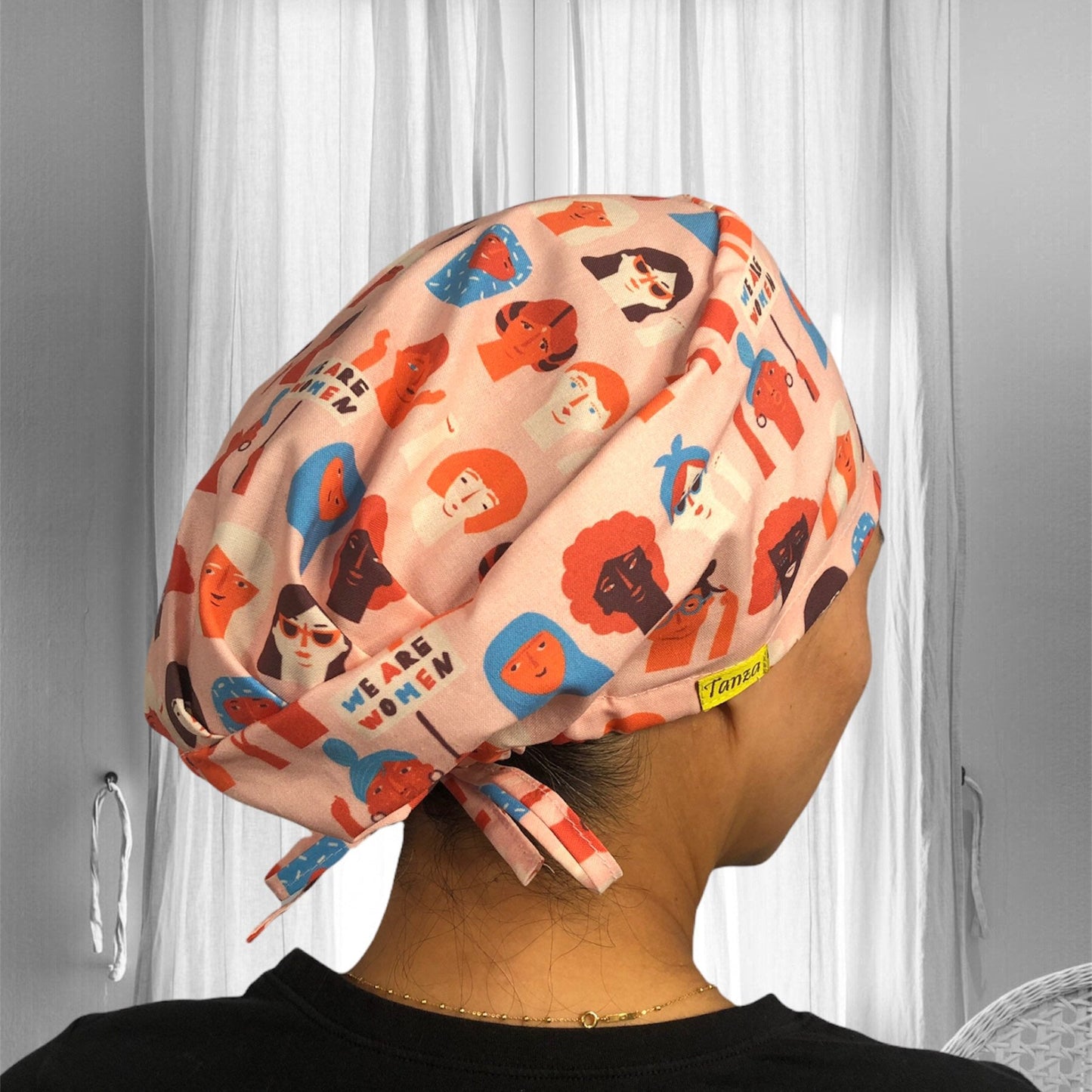 A colorful scrub cap featuring a variety of illustrated faces in vibrant shades of orange, blue, and black. The illustrations depict diverse women, each with unique hairstyles and accessories, celebrating empowerment. The text "WE ARE WOMEN" is prominently displayed on the fabric.