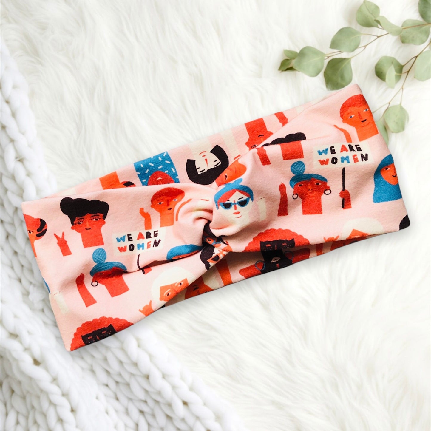 A colorful headband featuring a variety of illustrated faces in vibrant shades of orange, blue, and black. The illustrations depict diverse women, each with unique hairstyles and accessories, celebrating empowerment. The headband has a twisted knot design in the center, and the text "WE ARE WOMEN" is prominently displayed on the fabric.