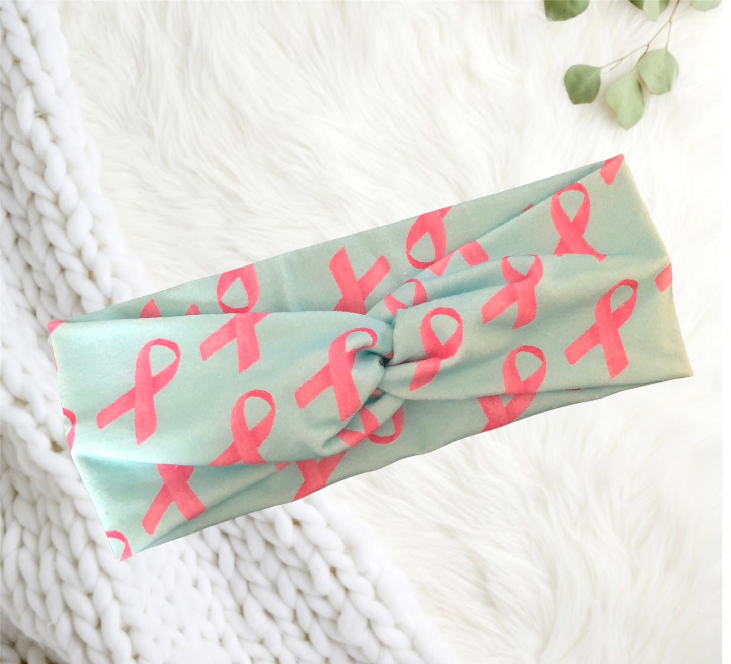 A stylish headband with a turquoise background featuring a pattern of pink ribbons relating it to breast cancer awareness. The headband is twisted in the center, adding an elegant touch.