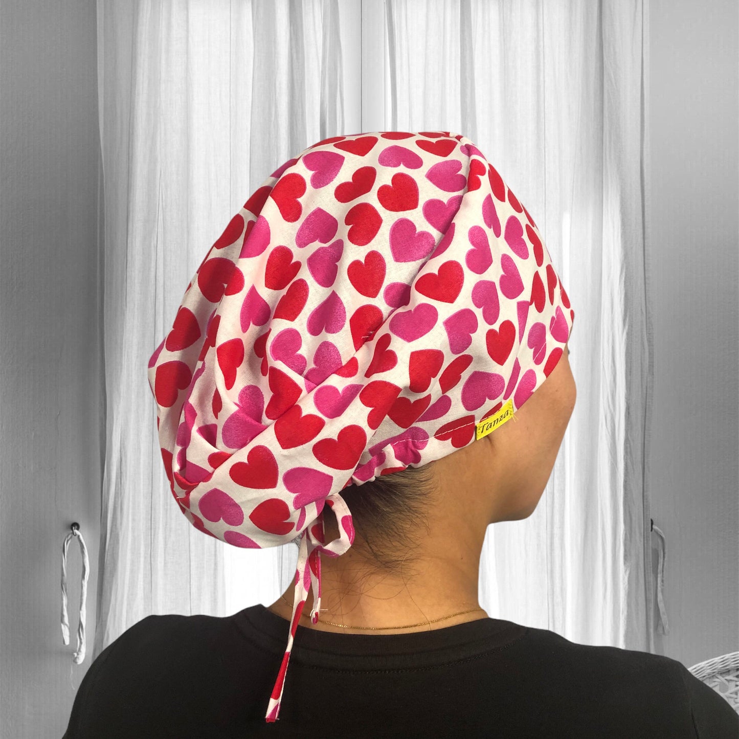 A person wearing a stylish scrub cap adorned with a pattern of red and pink hearts, set against a light background.