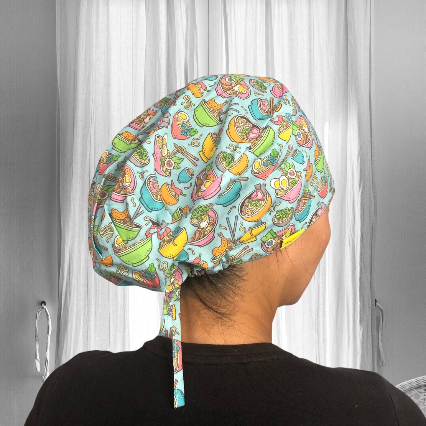 A person wearing a colorful scrub cap with a pattern featuring various types of Asian cuisine dishes, including bowls of noodles, sushi, and dumplings.