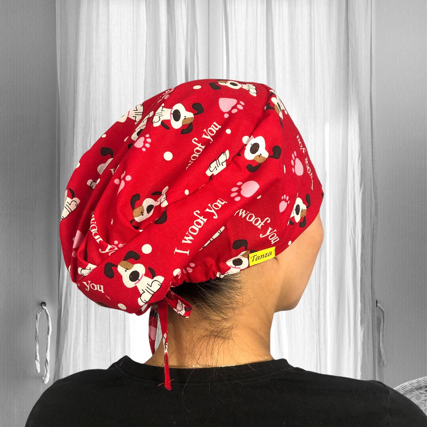 A person wearing a scrub cap featuring a playful dog pattern. The cap is adorned with various cartoon-style dog faces in different colors and expressions, set against a dark background. The fabric is decorated with small bones and paw prints, adding to the whimsical theme.