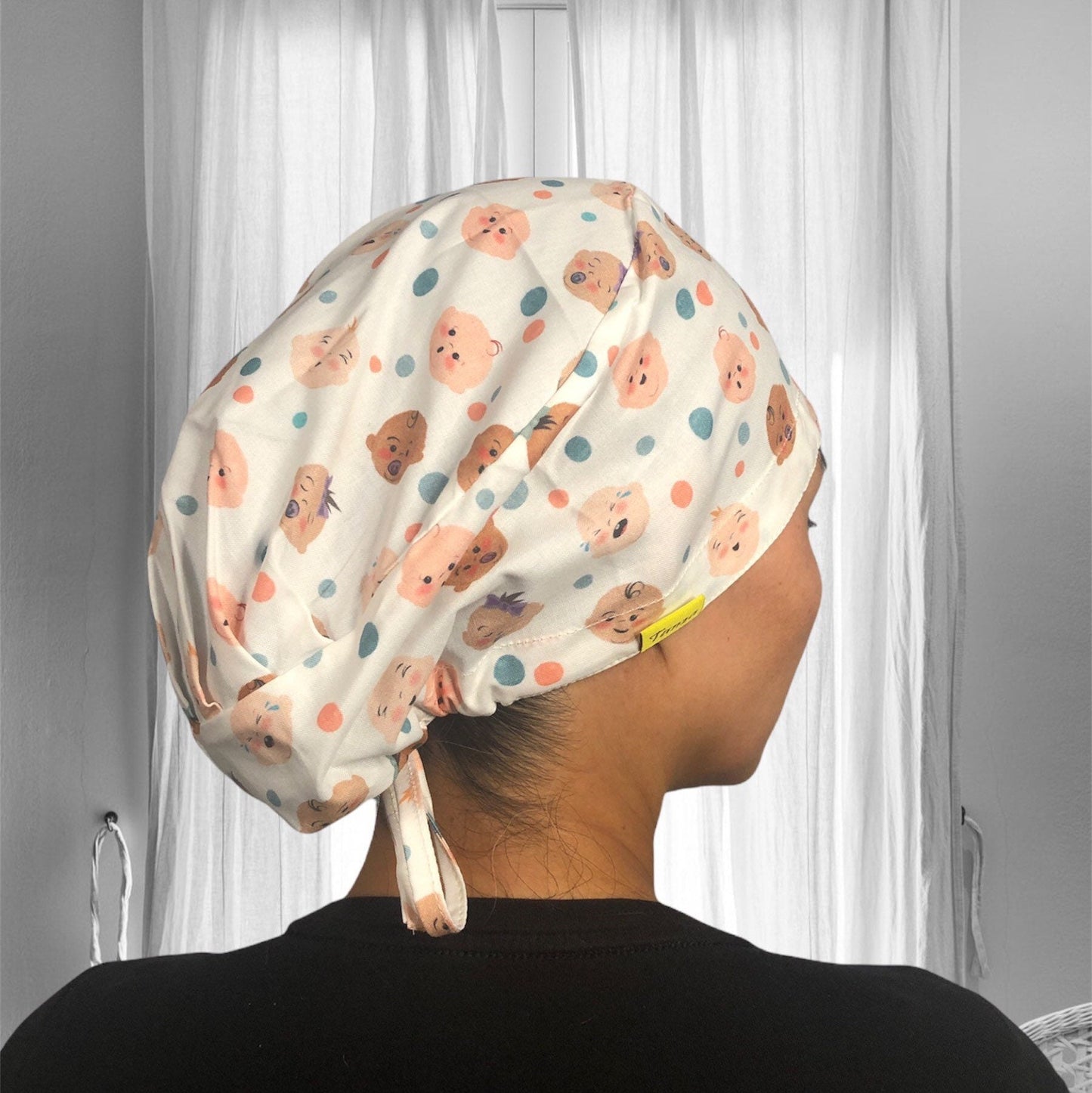 A scrub cap with a playful pattern of various cartoon baby faces and polka dots in soft pastel colors.