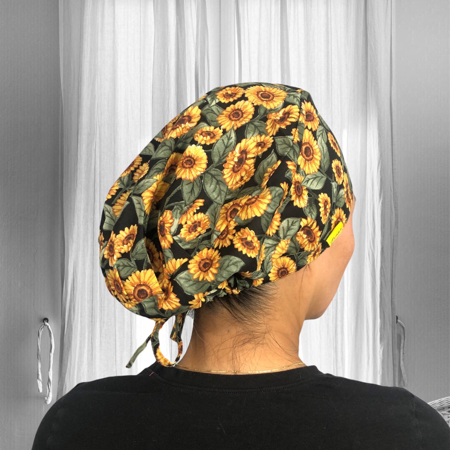 A person wearing a scrub cap adorned with a vibrant sunflower pattern. The cap is predominantly black, which makes the bright yellow sunflowers stand out. The design includes detailed green leaves surrounding the flowers.