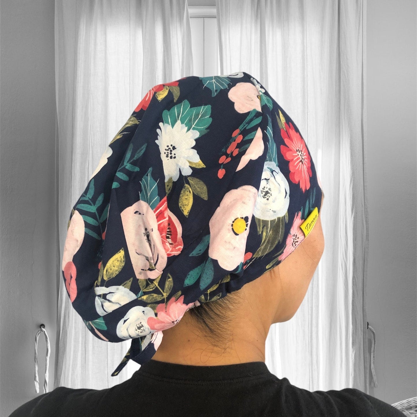 A person wearing a stylish floral scrub cap with a vibrant pattern of flowers in various colors. The cap has a dark background.