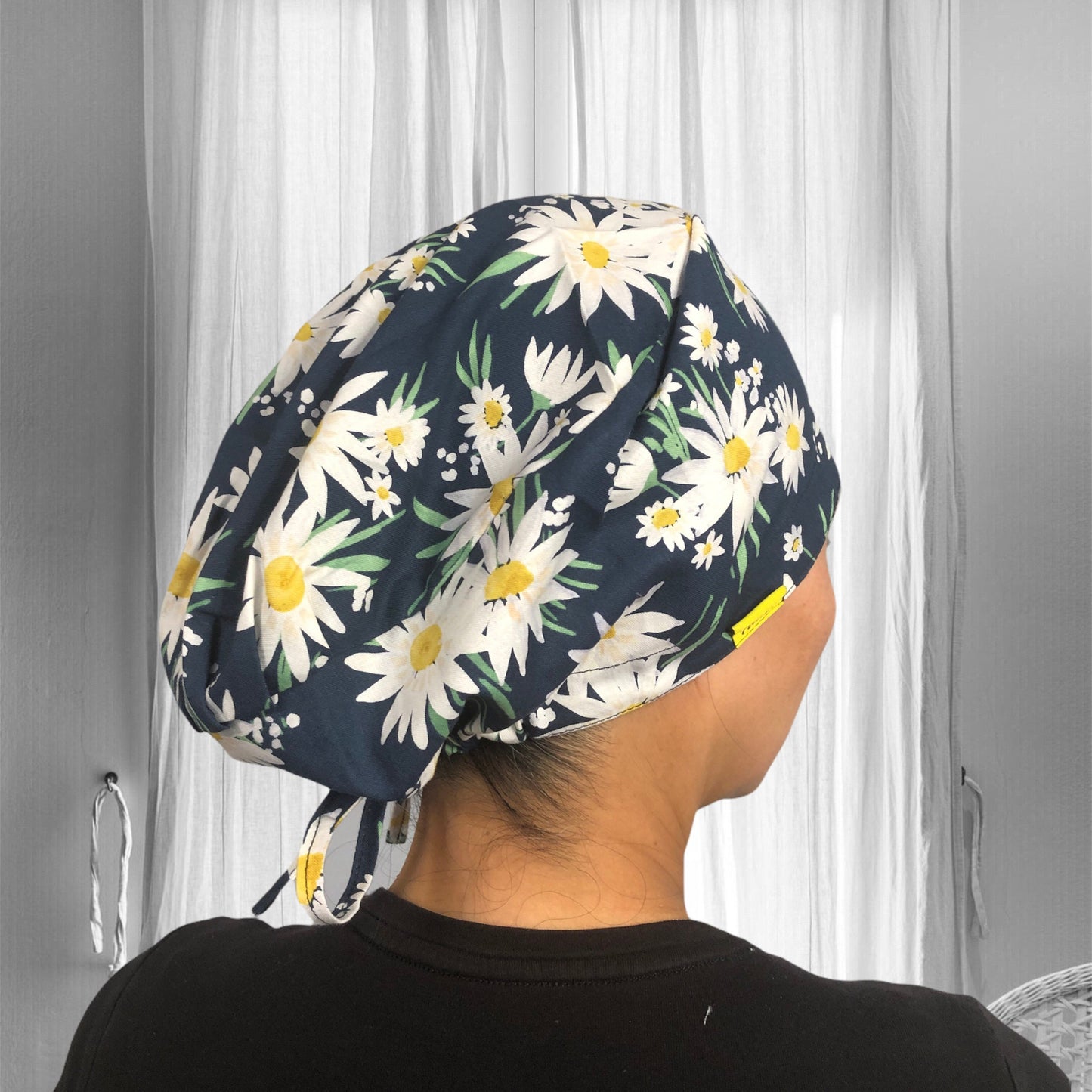 A person wearing a floral scrub cap, featuring a navy blue background adorned with large, white daisies and green leaves.
