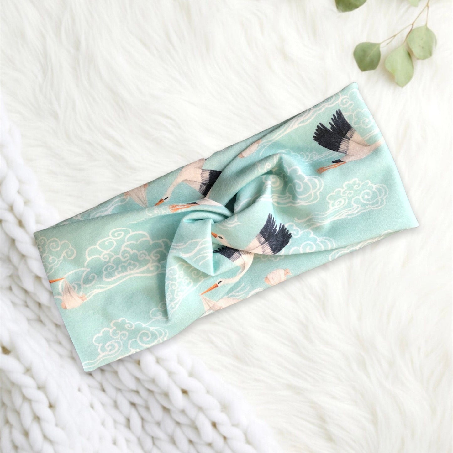 A stylish headband made from soft fabric in a delicate mint green shade, featuring an elegant pattern of cranes soaring among swirling clouds. The headband is artfully knotted in the center.