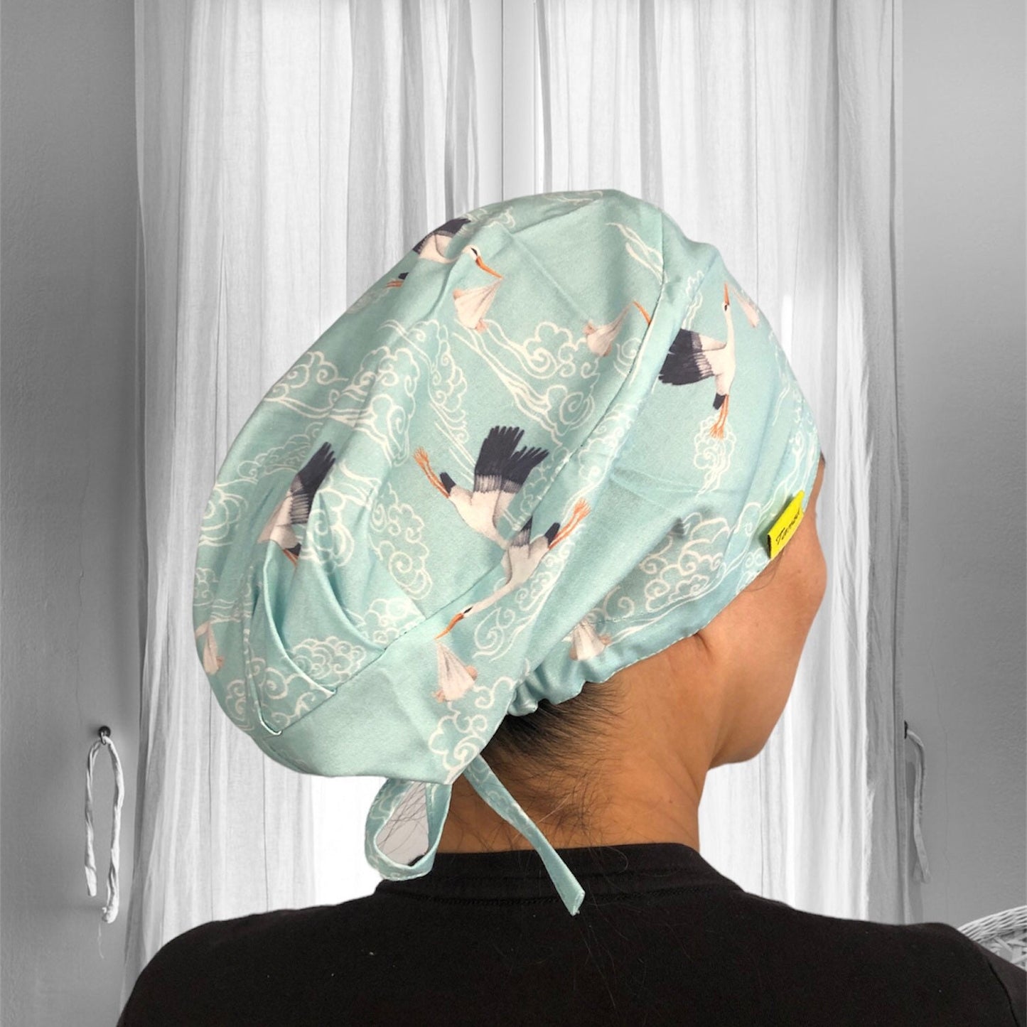 A stylish scrub cap made from soft fabric in a delicate mint green shade, featuring an elegant pattern of cranes soaring among swirling clouds.