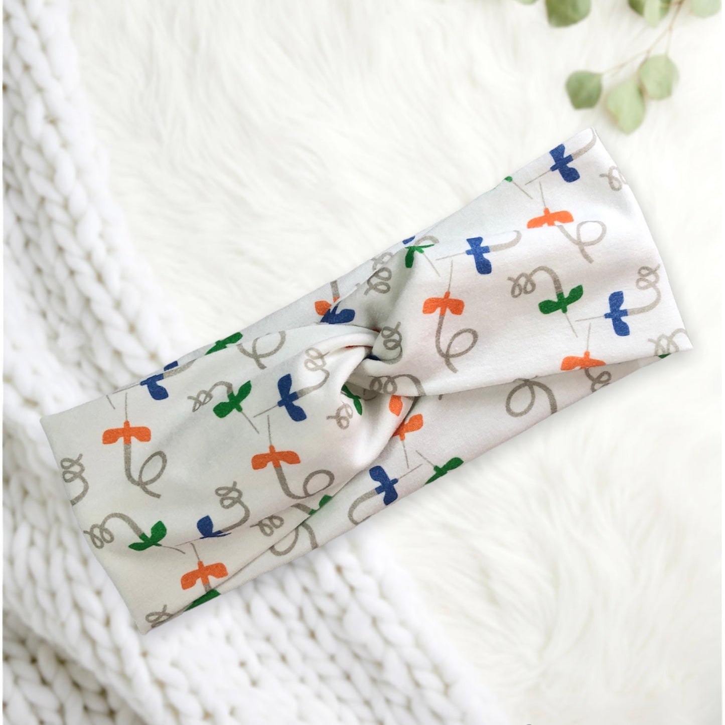 A soft fabric headband featuring a playful pattern of multicolored needles in orange, green, and blue on a light background. The headband is styled with a central twist.