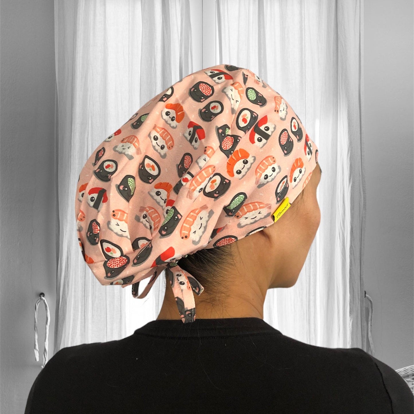 A person wearing a pink scrub cap adorned with a playful sushi print, featuring various sushi rolls and pieces of nigiri.