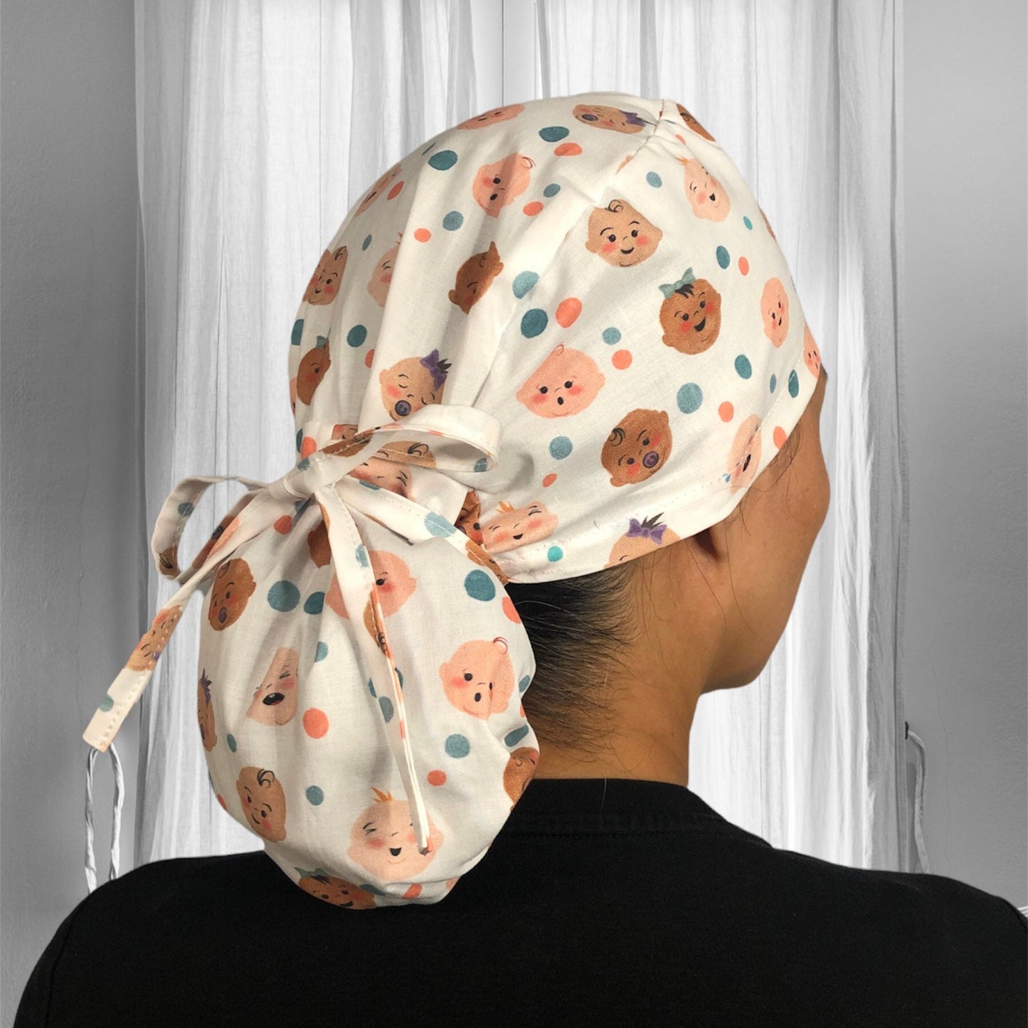 A patterned scrub cap featuring cute, cartoon-style baby faces of various expressions and hairdos in pastel colors.