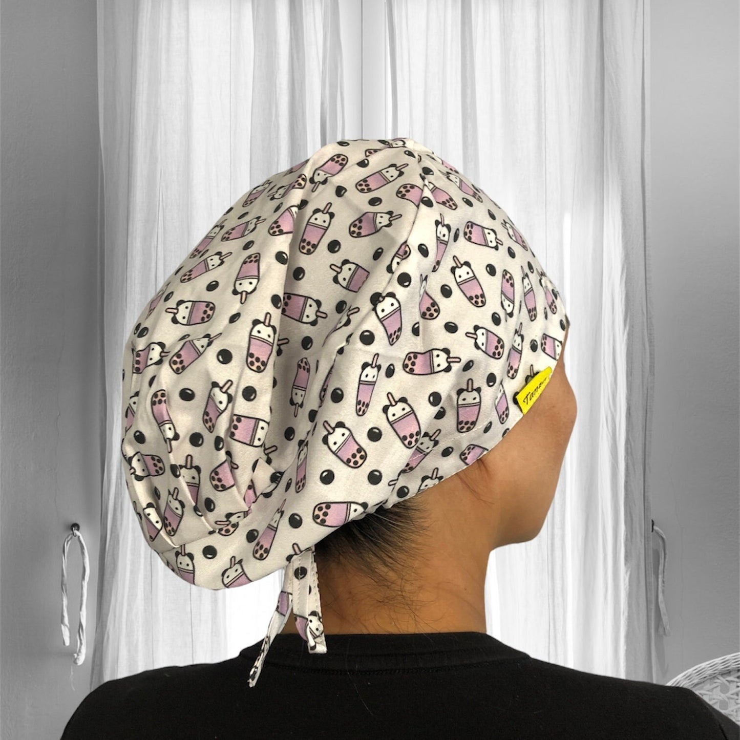 A person wearing a stylish scrub cap with a playful pattern of pink and white bubble tea cups and black bubbles.