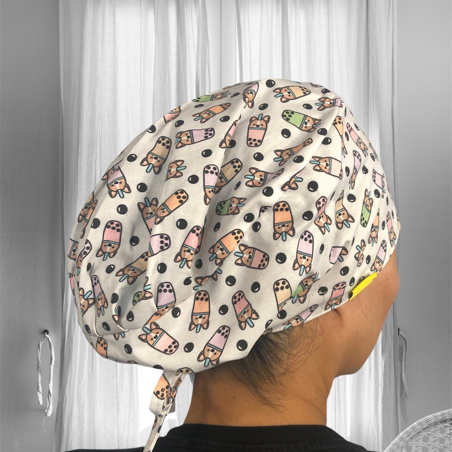 A person wearing a fabric cap with a playful pattern. The cap is adorned with cartoon-like illustrations of corgi inside of a milktea in various colors, including green, pink, and brown, scattered across a white background with black bobas.